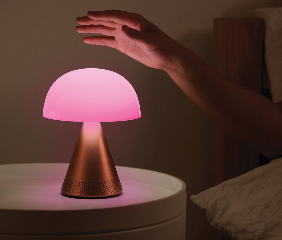 Eine Hand reicht nach der eingeschalteten goldenen LEXON Akku-Tischleuchte »Mina L Audio« auf einem Nachttisch neben einem Bett.