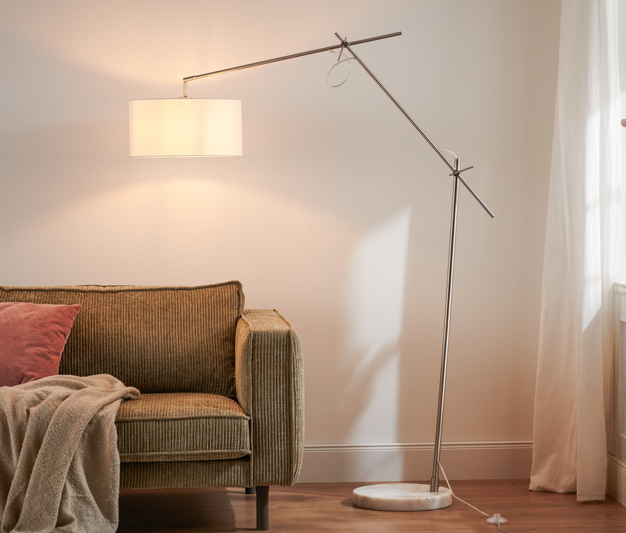 Ein braunes Sofa mit einem rosa Kissen und einer beigen Decke, daneben steht eine Stehleuchte mit Textilschirm.