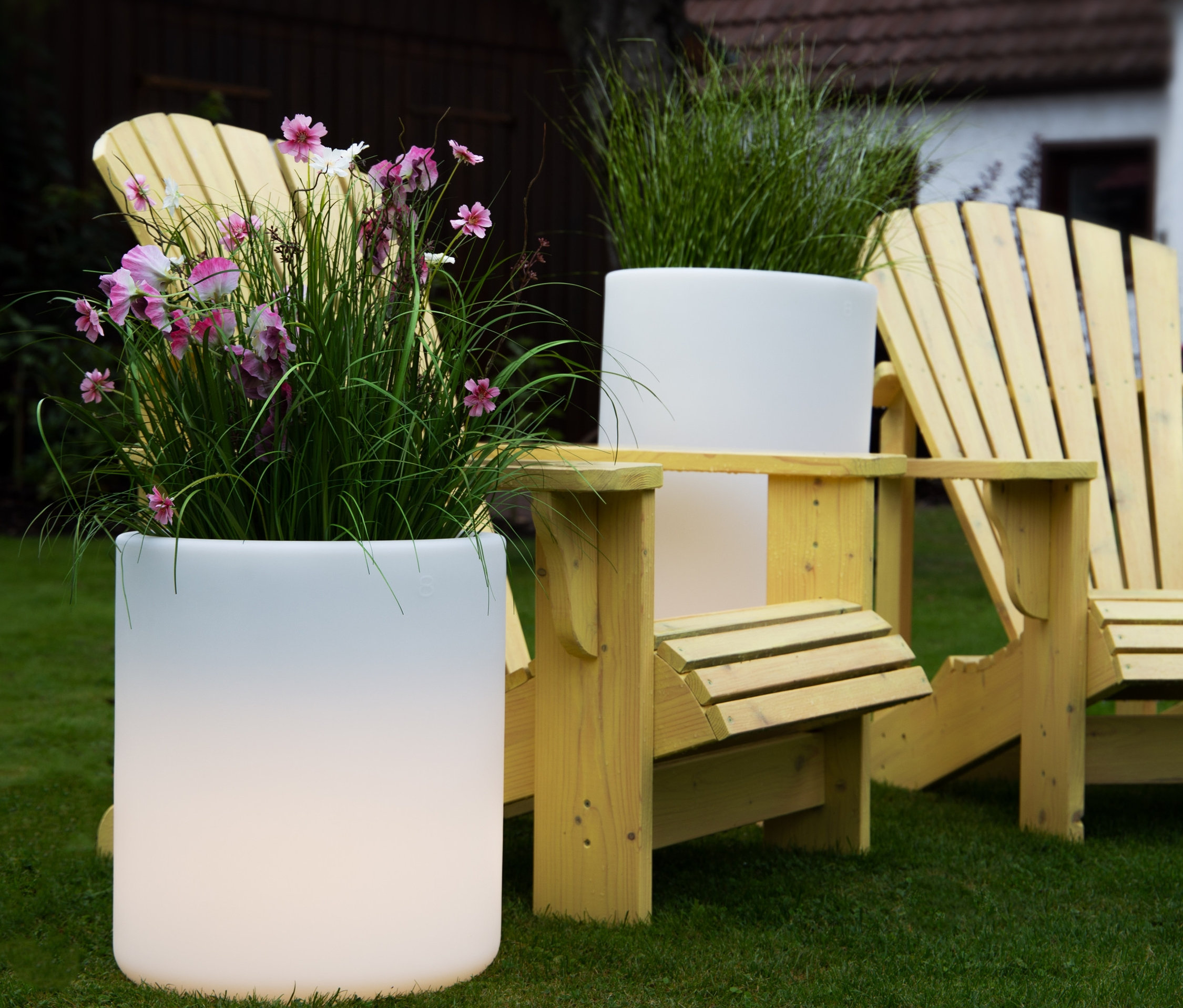 Zwei gelbe Holzstühle stehen auf einer Rasenfläche neben zwei weißen Pflanzgefäßen, eines mit rosa Blumen. Eine 8 seasons design LED-Leuchte ist auch zu sehen.