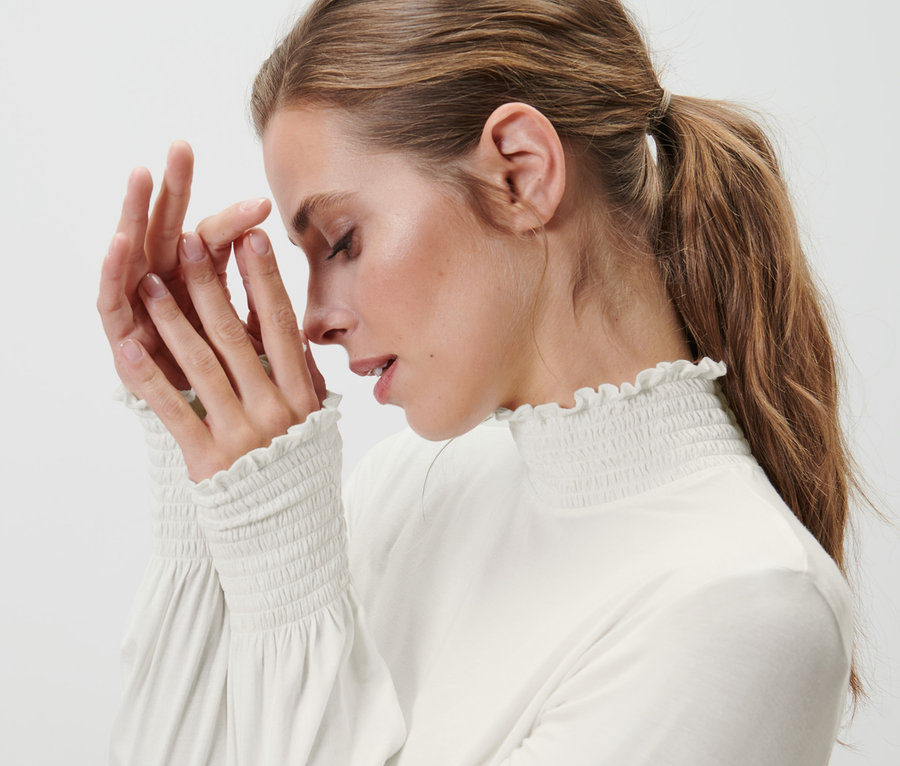 Frau berührt mit geschlossenen Augen ihr Gesicht. Sie trägt ein cremefarbenes Blusenshirt mit Stehkragen.