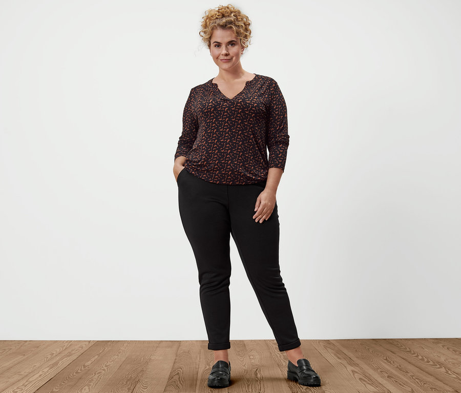 Eine Frau posiert in Joggpants, Leder-Loafern und einem bedruckten Blusenshirt.