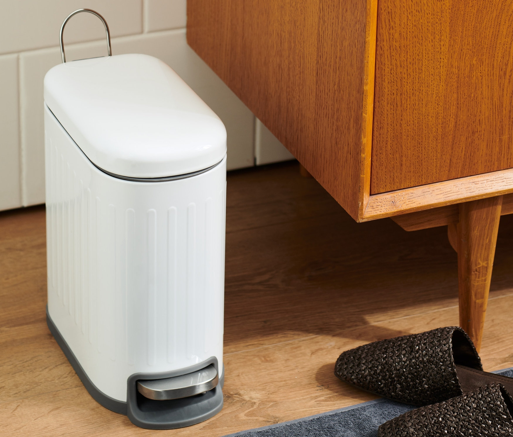 Im Badezimmer steht ein weißer Abfalleimer neben einem braunen Holzschrank und Hausschuhen auf einem grauen Teppich.