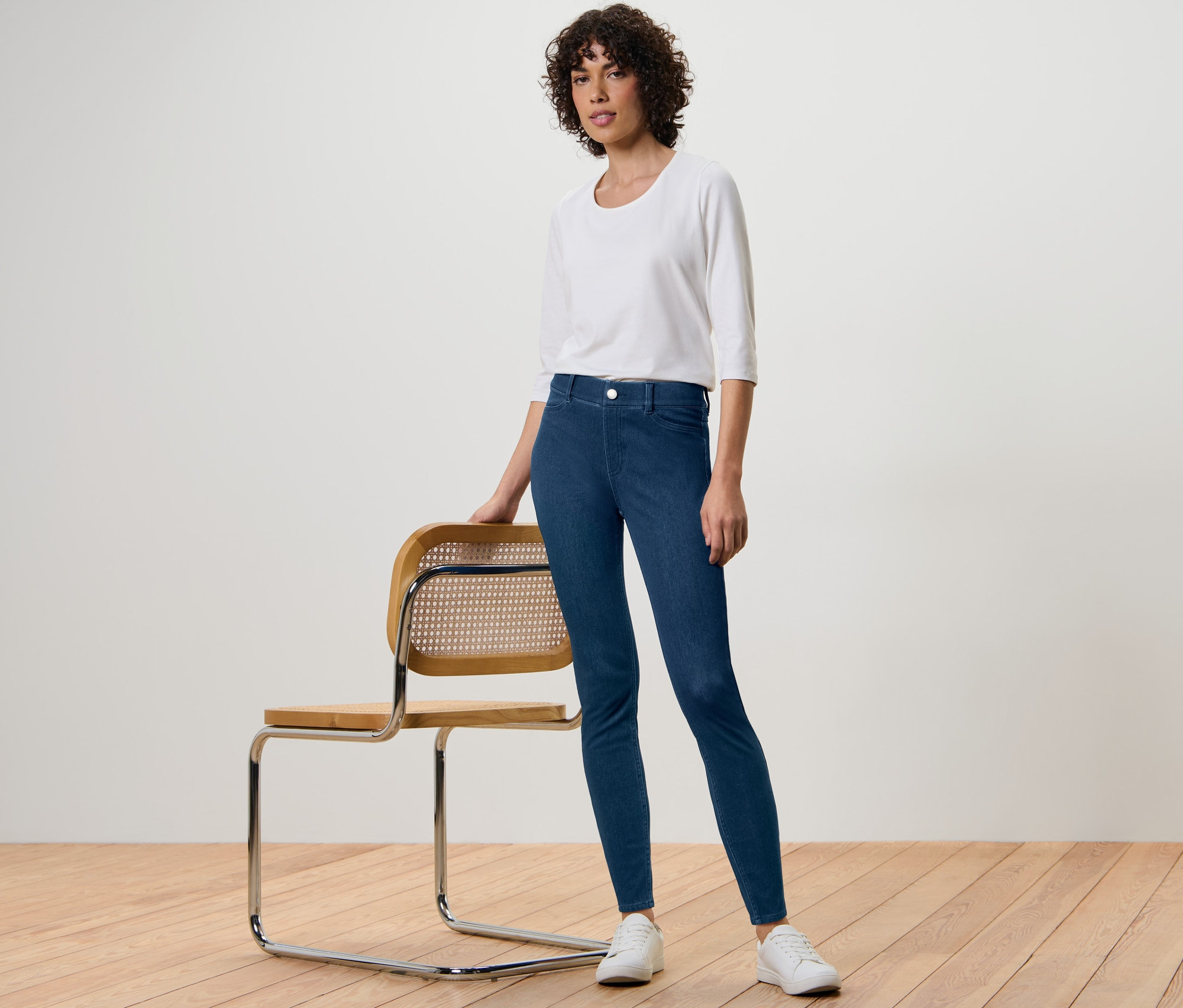 Frau posiert neben einem Stuhl und trägt Jeggings, eine weiße Bluse und Turnschuhe.