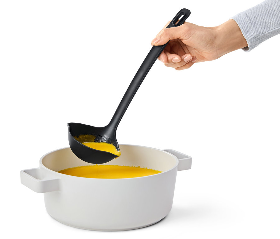 Eine Hand hält eine schwarze Multifunktions-Schöpfkelle über einen Topf mit orangefarbener Flüssigkeit.