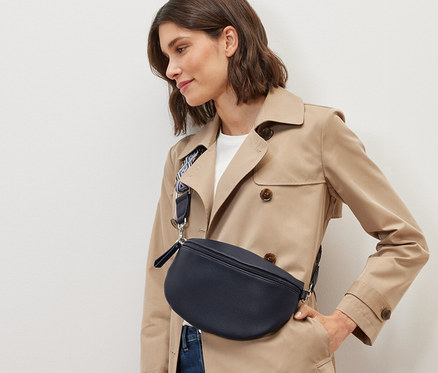 Frau im beigen Trenchcoat und Jeans posiert mit Crossbody-Tasche.