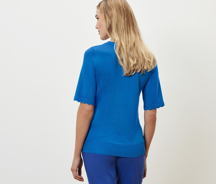 Blonde Frau steht mit dem Rücken zur Kamera und trägt einen kobaltblauen Feinstrickpullover und blaue Hosen.