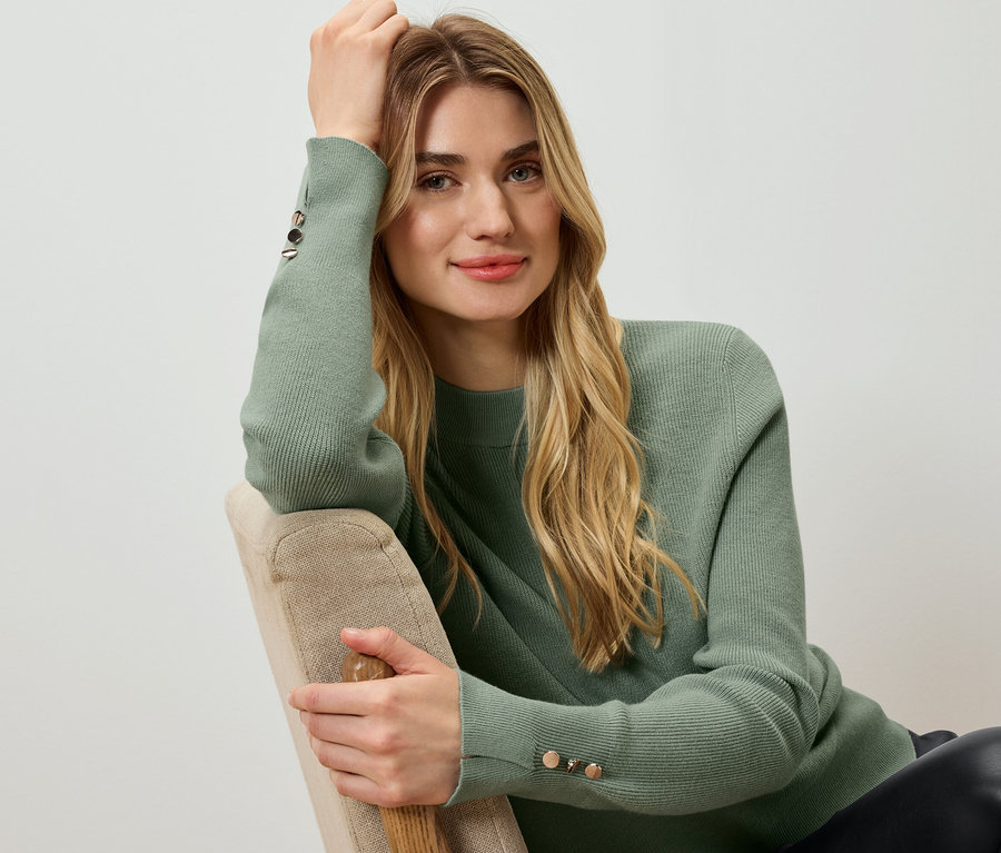 Frau sitzt, stützt ihren Kopf auf ihre Hand und trägt einen grünen Pullover.