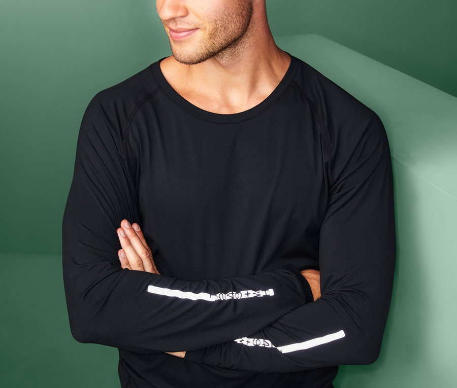 Ein Mann mit verschränkten Armen trägt eine schwarze Reflektor-Sportcap und ein schwarzes Langarmshirt mit reflektierenden Details.