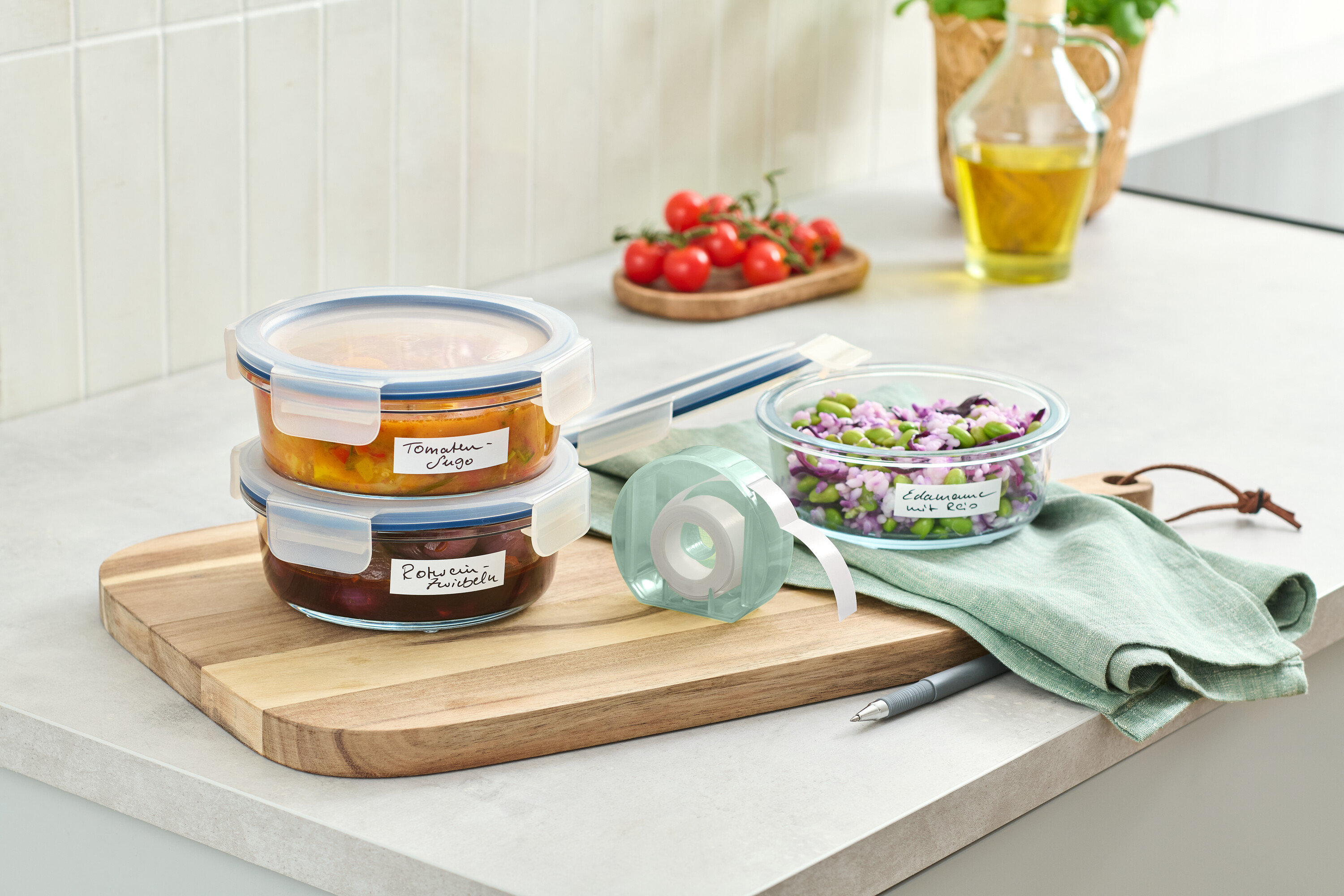 Zwei Glasbehälter mit Essen gestapelt auf einem Holzbrett neben einer Etikettenrolle.