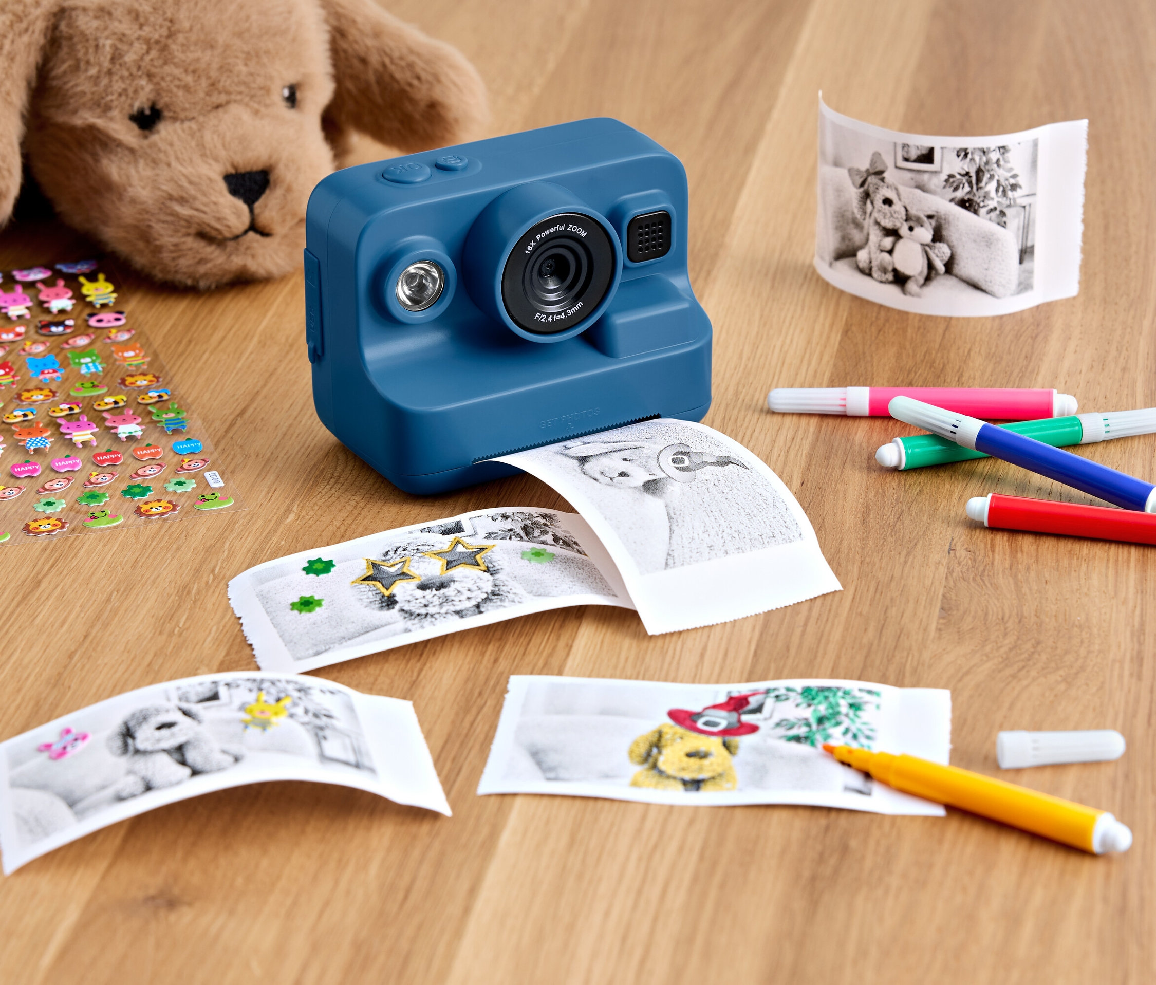 Eine blaue Sofortbildkamera liegt auf einem Holztisch mit einem Plüschteddy, einem Bogen Aufkleber, ausgedruckten Fotos und bunten Markern.