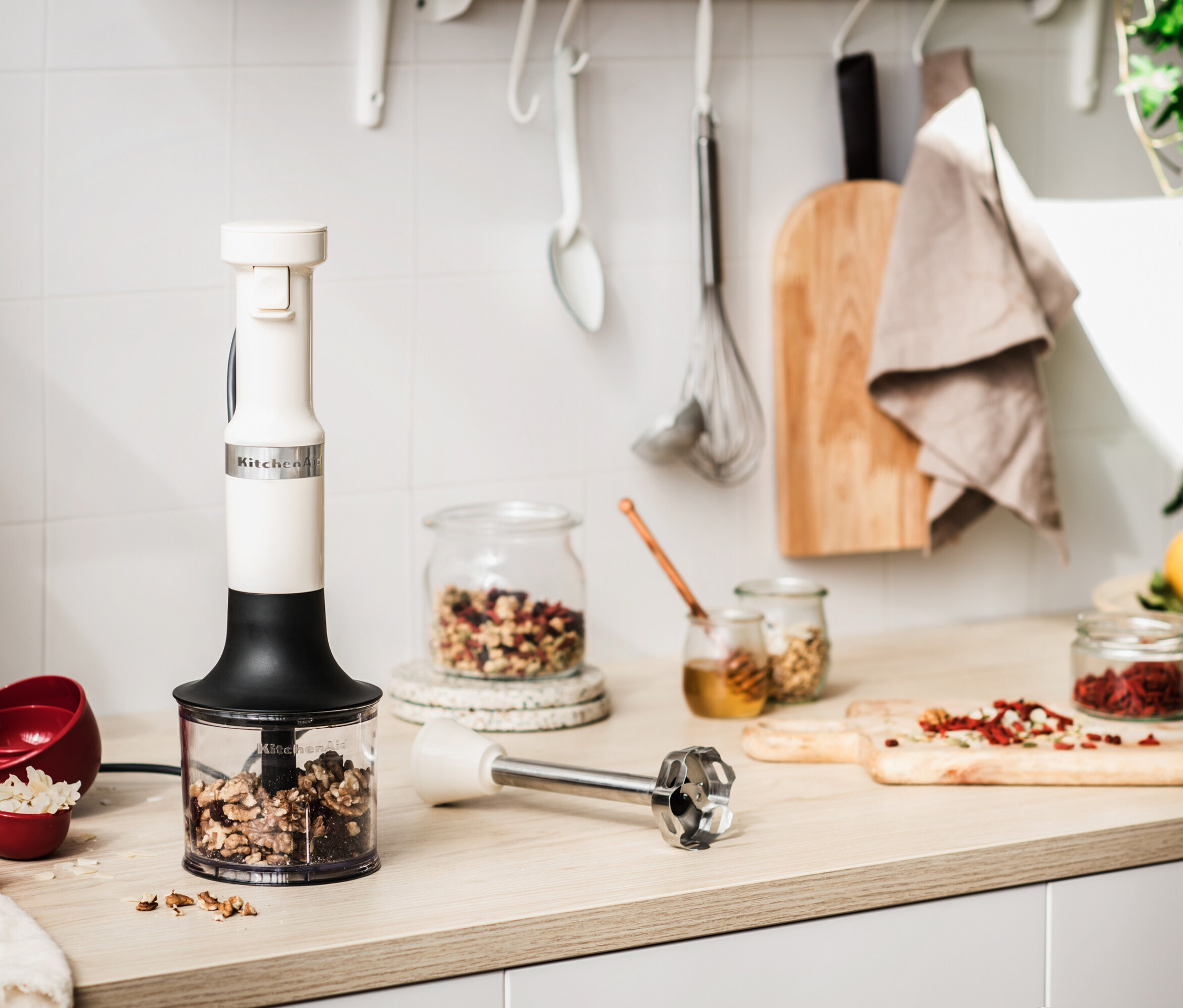 Ein Stabmixer mit einem Behälter voller Nüsse steht auf einer Küchentheke neben Gläsern mit Nüssen und Honig.