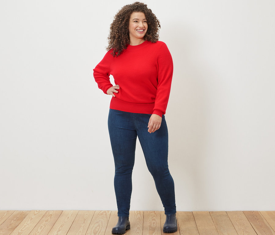 Frau posiert in rotem Feinstrickpullover und High-Waist-Denim-Leggings.