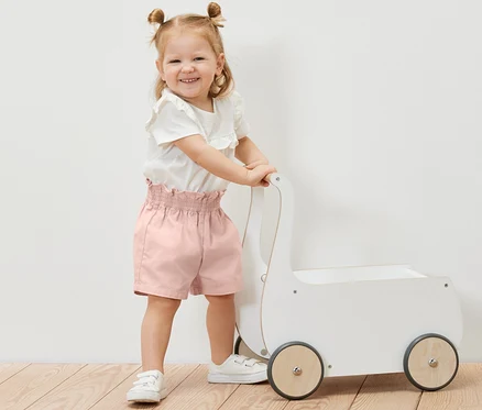 Lächelndes kleines Mädchen schiebt einen weißen Spielzeugwagen und trägt eine Kinder-Paperbag-Shorts.