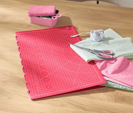 Eine rosa faltbare Schneide- und Bastelunterlage liegt auf einem Tisch neben einer rosa Box mit einer Schere, Stoffen und einem Maßband.