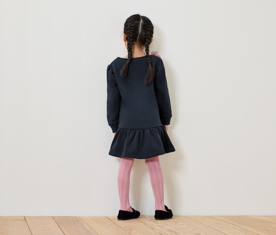 Ein Mädchen mit Zöpfen steht mit dem Rücken zur Kamera und trägt ein blaues Kleid, eine rosa gemusterte Strumpfhose und schwarze Schuhe.