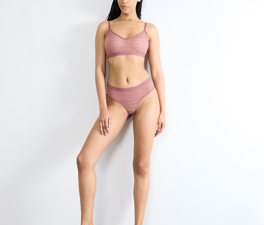 Barfuß stehende Frau vor einer weißen Wand, die einen pinkfarbenen sloggi FREE Evolve Hipster Lace in Date Night Pink und einen passenden Bralette trägt.