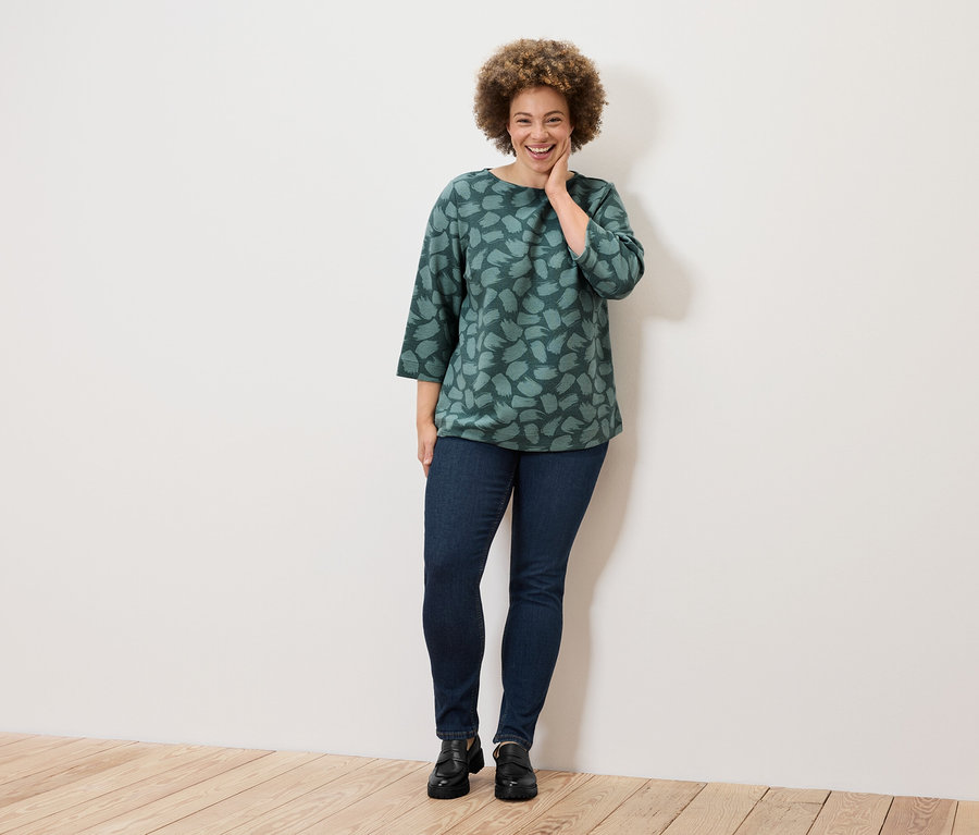 Eine lächelnde Frau mit lockigem Haar posiert in einem grünen Top mit Muster, Jeans und schwarzen Schuhen.