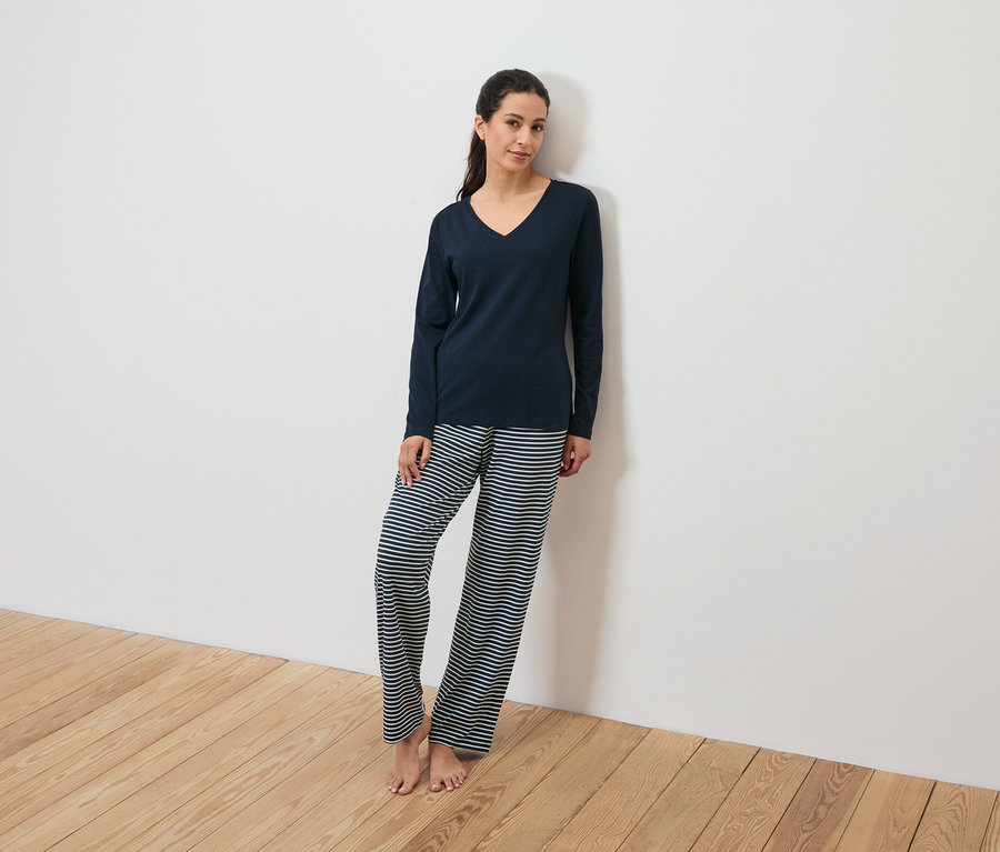 Eine Frau in einem marineblauen Pyjamaoberteil und einer gestreiften Hose lehnt an einer weißen Wand.