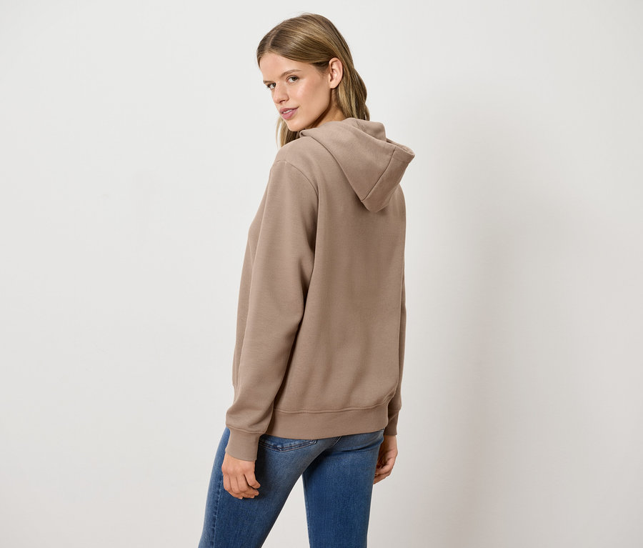 Frau blickt über die Schulter und trägt einen braunen Kapuzenpullover und blaue Jeans.