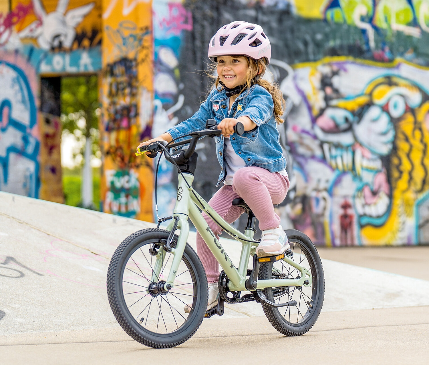 Ein junges Mädchen mit rosa Helm fährt ein mintgrünes PUKY LS-PRO 16 Fahrrad vor einer Wand mit Graffiti.