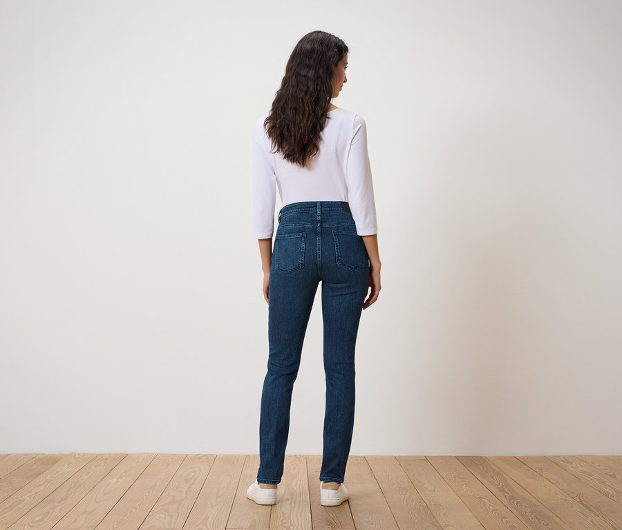 Eine Frau mit dunklem, gewelltem Haar steht mit dem Rücken zum Betrachter, sie trägt ein weißes Dreiviertelarm-Shirt und dunkelblaue Jeans.