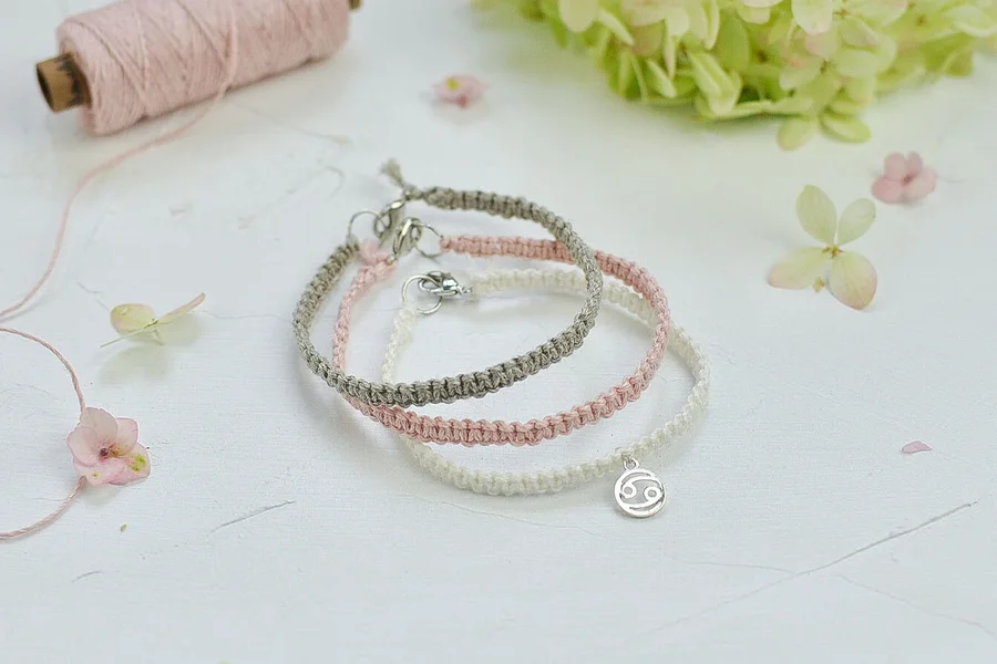 Drei geflochtene Armbänder in Grau, Rosa und Weiß liegen auf einer weißen Oberfläche. Eine weiße Blume und eine rosa Schnur sind im Hintergrund.