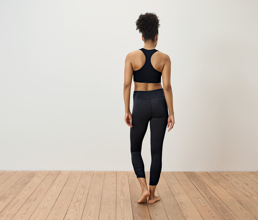 Frau in schwarzer Sportbekleidung steht mit dem Rücken zur Kamera auf einem Holzboden.