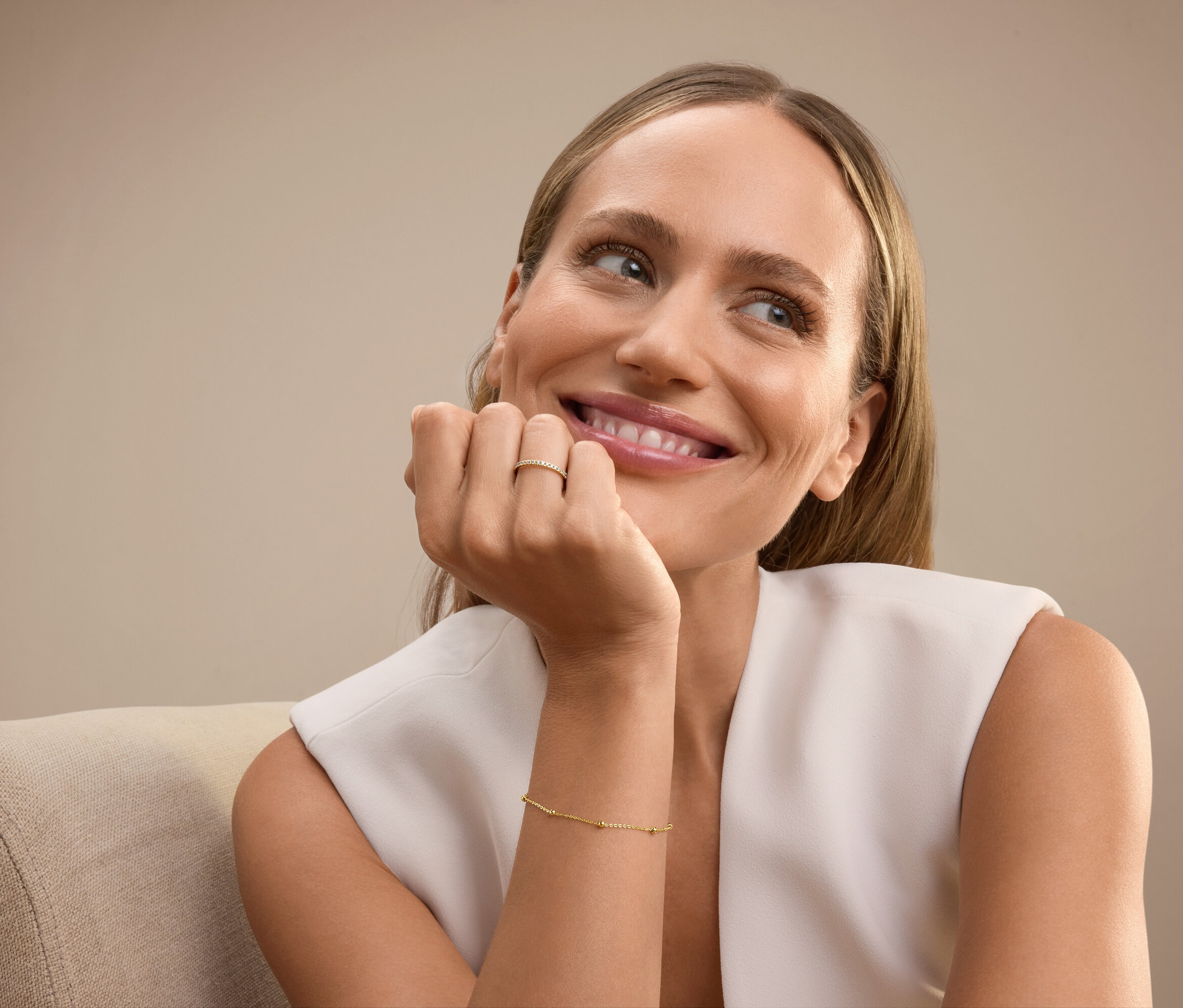 Lächelnde Frau sitzt auf einem Stuhl, trägt ein goldenes Armband am Arm und einen Ring am Ringfinger.