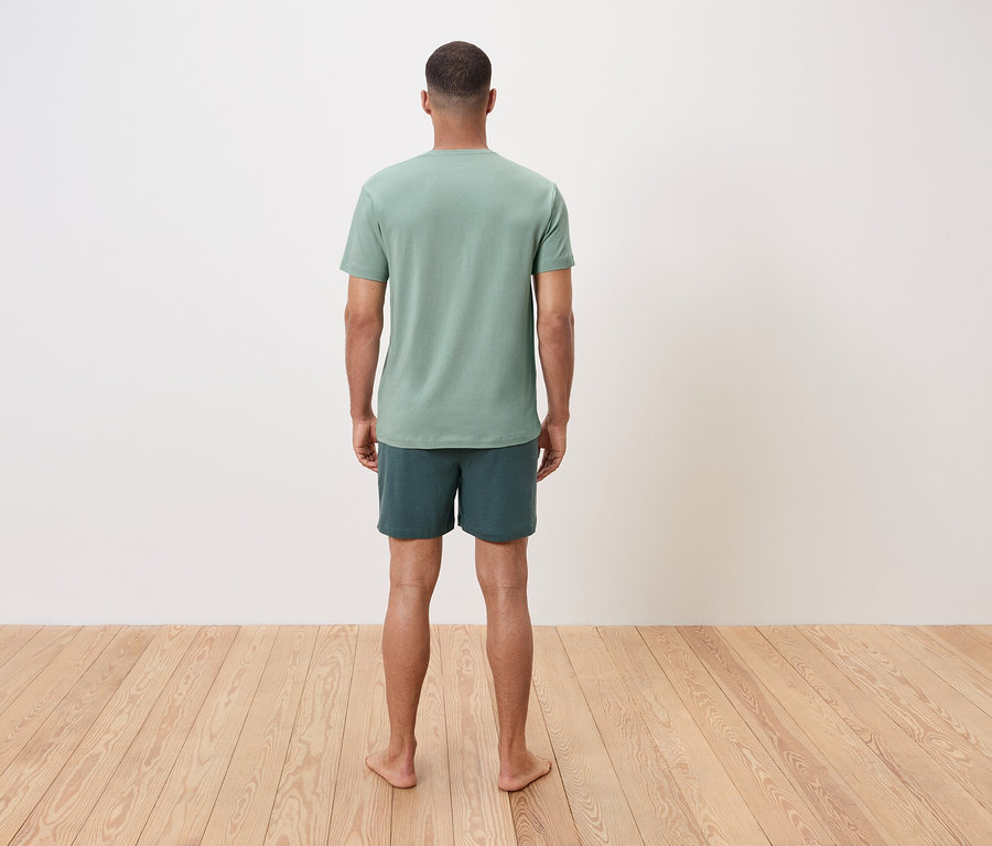 Ein Mann steht barfuß mit dem Rücken zum Betrachter, er trägt ein grünes T-Shirt und Shorts.