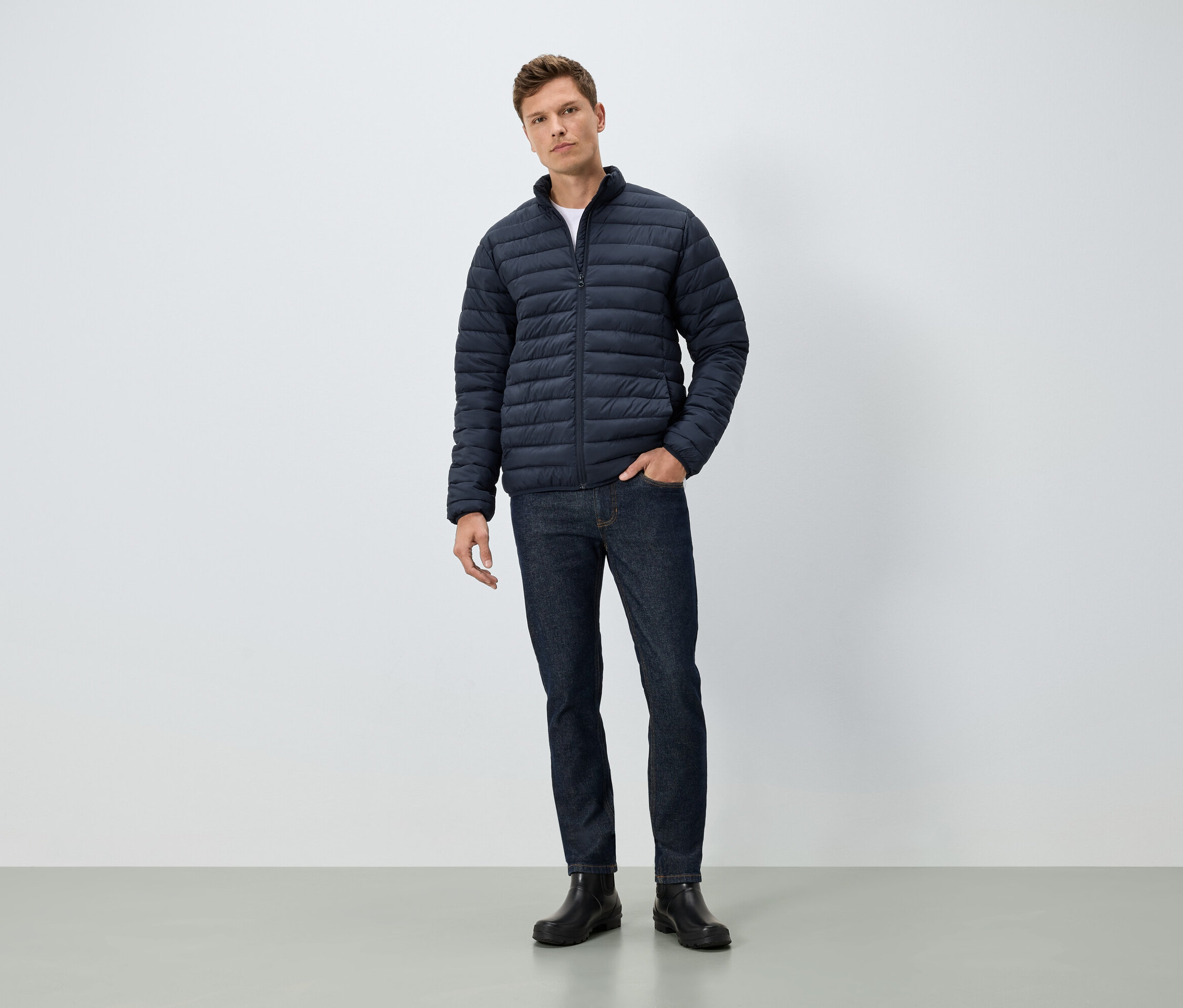 Ein Mann steht mit einer Hand in der Tasche. Er trägt eine blaue Jacke, Jeans und schwarze Stiefel.