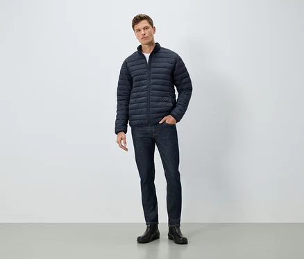 Ein Mann steht mit einer Hand in der Tasche. Er trägt eine blaue Jacke, Jeans und schwarze Stiefel.