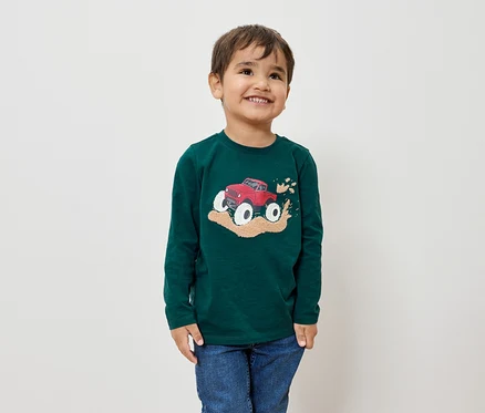 Ein lächelnder Junge trägt ein grünes Langarmshirt mit einem roten Monstertruck-Bild und blaue Jeans.