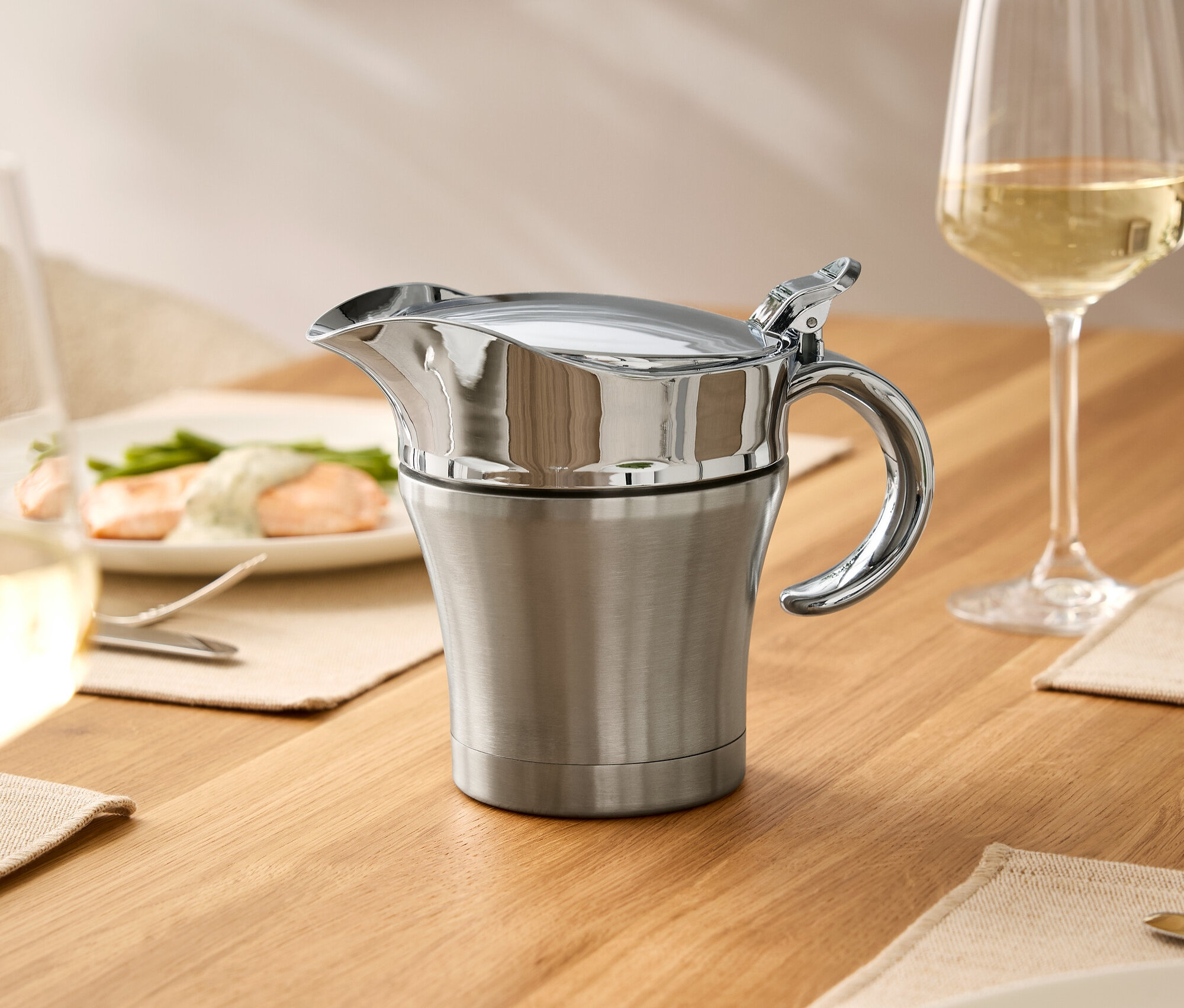 Auf einem Holztisch stehen eine Edelstahl-Soßenschüssel mit Deckel, ein Weinglas und Teller mit Speisen.