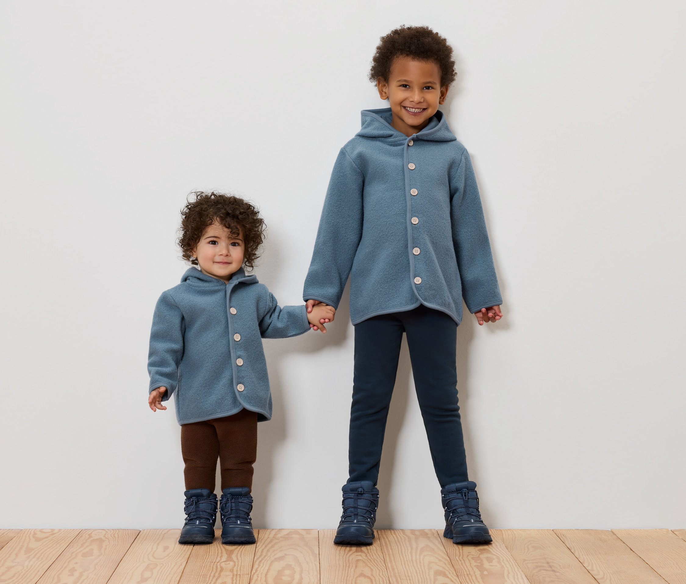 Zwei Kinder halten Händchen. Beide tragen blaue Mäntel und Stiefel.