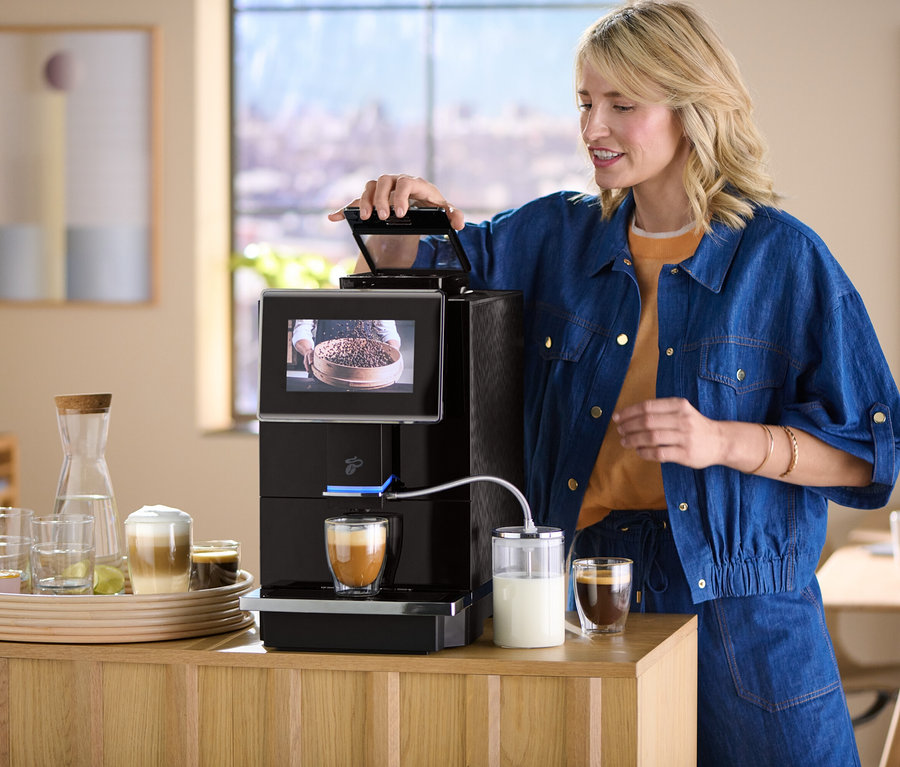 Blonde Frau öffnet den Deckel einer Kaffeemaschine, auf dem Tisch ein Tablett mit Gläsern.