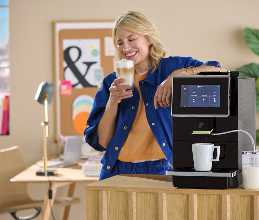 Eine lächelnde Frau hält ein Glas Milchkaffee neben einer Kaffeemaschine.