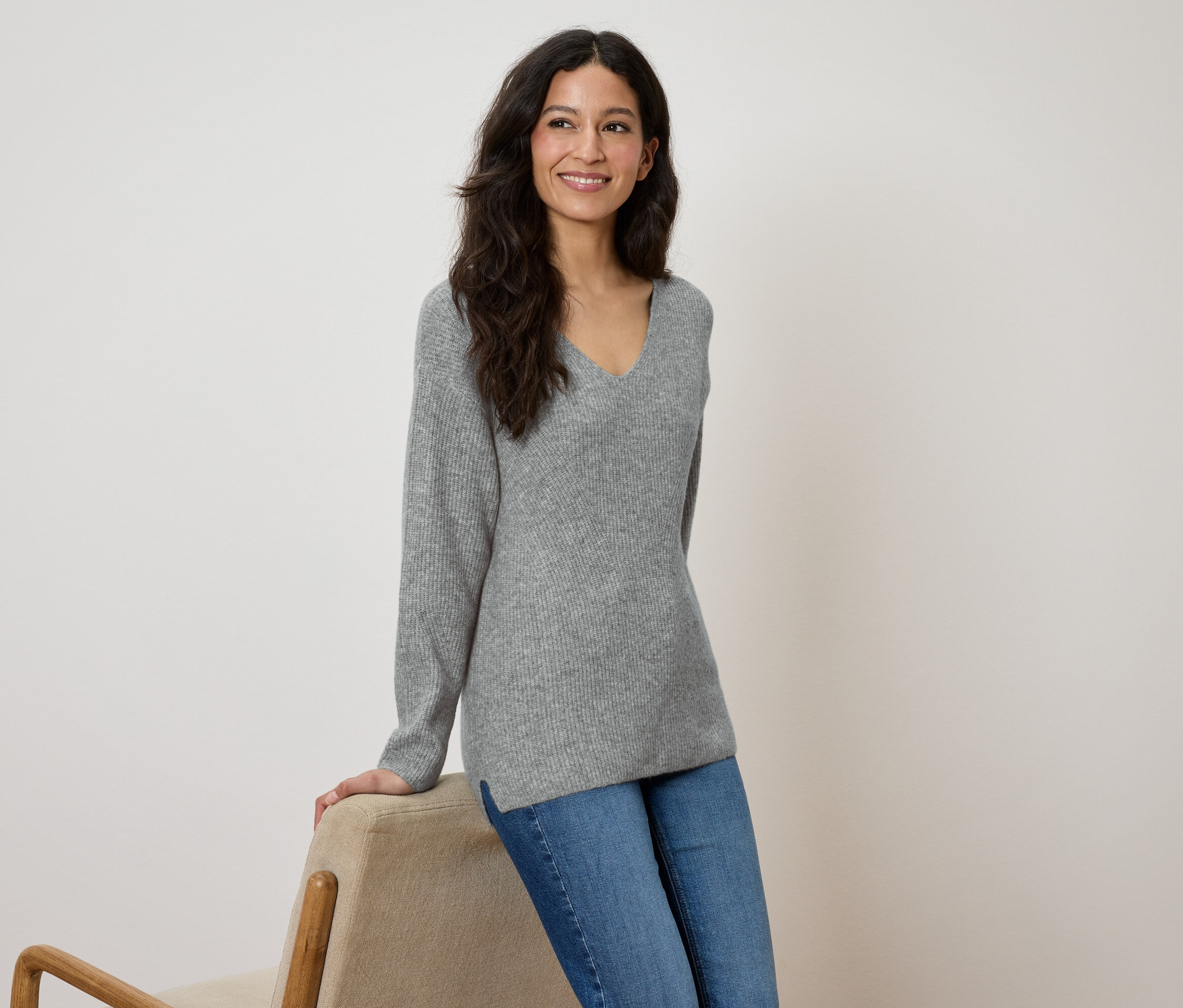 Eine Frau lächelt und lehnt sich an einen beigen Stuhl, sie trägt einen grauen Pullover und blaue Jeans.