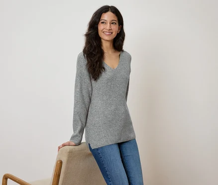 Eine Frau lächelt und lehnt sich an einen beigen Stuhl, sie trägt einen grauen Pullover und blaue Jeans.