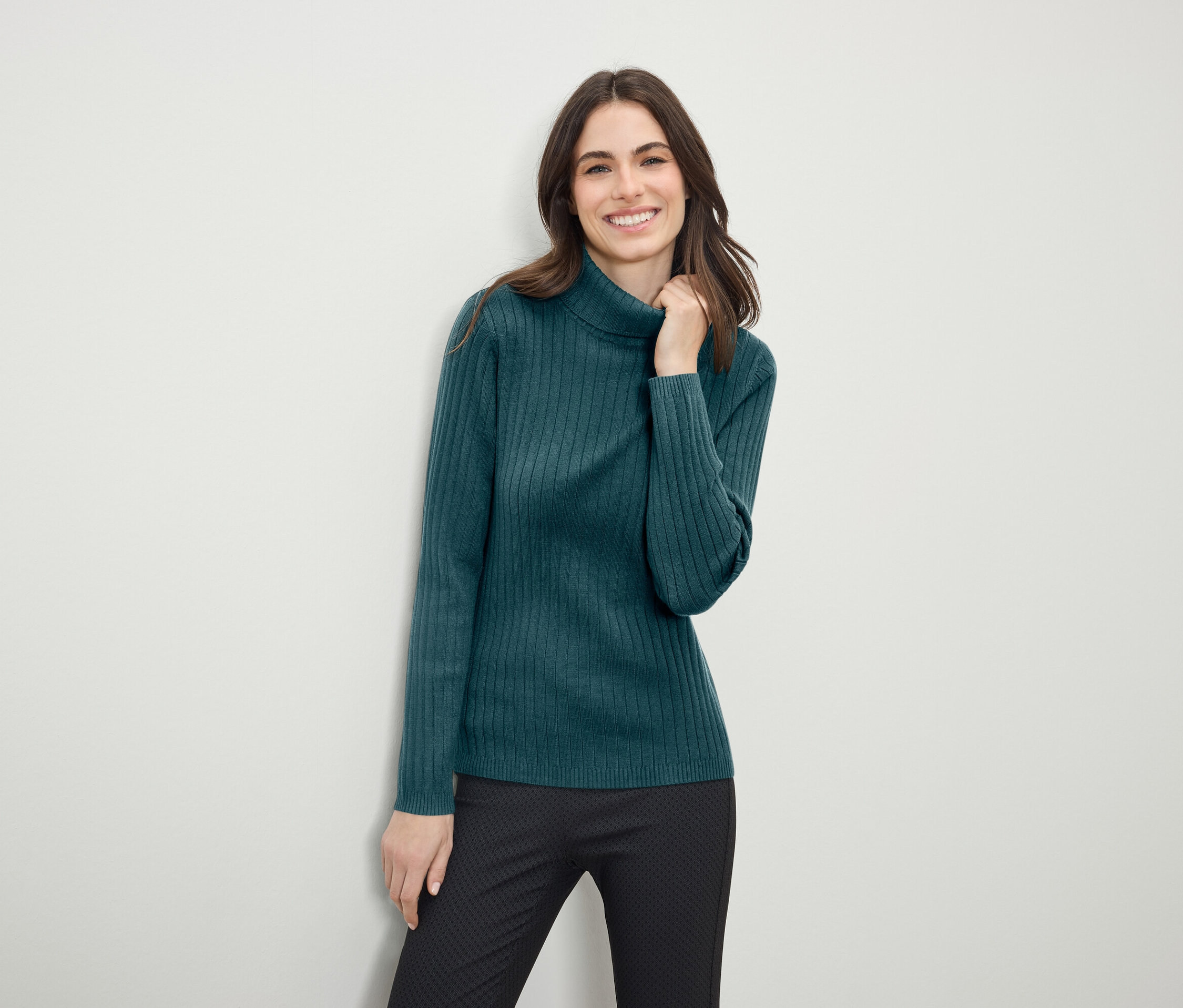 Eine lächelnde Frau posiert in einem grünen Rollkragenpullover und einer schwarzen Hose.