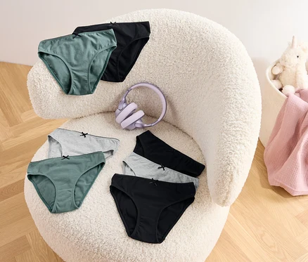 Sechs Slips in Grün, Grau und Schwarz und ein lila Kopfhörer liegen auf einem zotteligen Sessel. In einem Korb befinden sich eine rosa Decke und ein Plüscheinhorn.