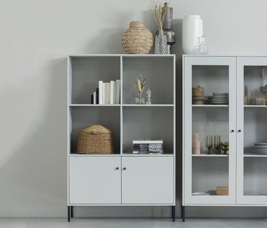 Ein graues Bücherregal mit sechs Fächern und zwei Türen steht neben einem Glasschrank.