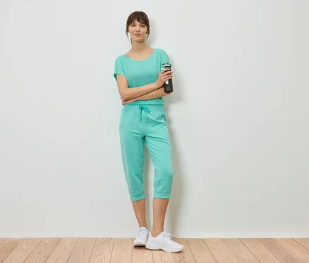 Frau in mintfarbenem Shirt und Caprihose steht mit verschränkten Armen und hält eine Wasserflasche.