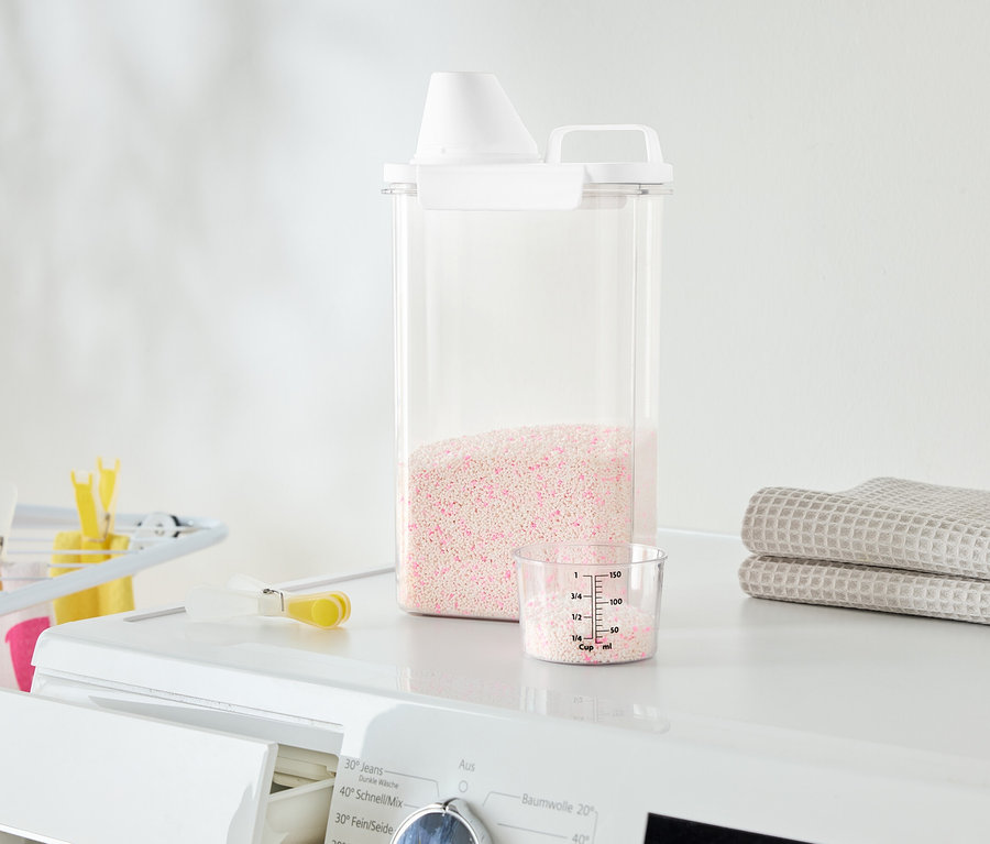 Auf einer Waschmaschine stehen ein durchsichtiger Behälter mit rosa Waschmittel, ein Messbecher und gefaltete Handtücher.