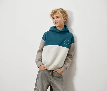 Ein blonder Junge lehnt an einer Wand und trägt einen blau-weiß-braunen Kapuzenpullover und graue Jeans.