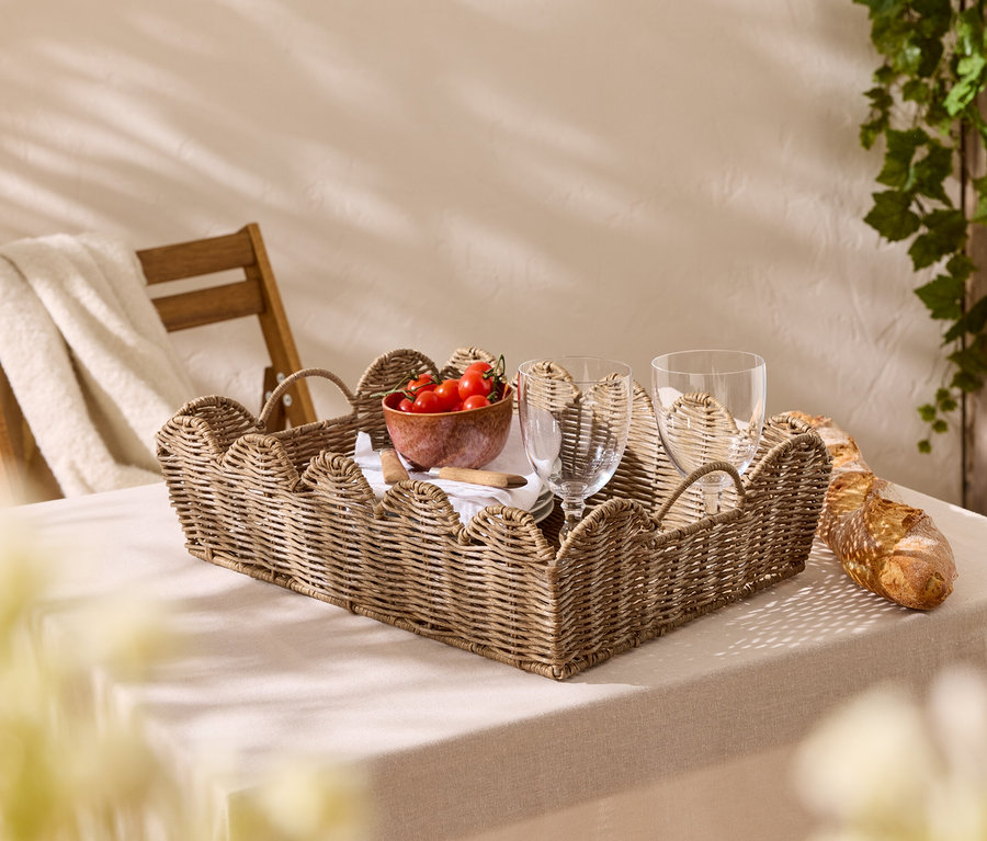 Auf einem Tisch mit Tischdecke steht ein geflochtenes Tablett mit Tomaten, Gläsern und Baguette.