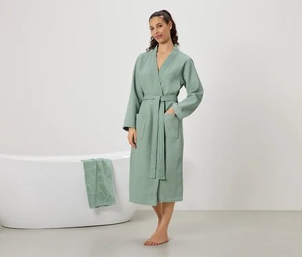Frau steht barfuß in einem mintgrünen Bademantel mit Gürtel und Taschen, eine Hand in der Tasche. Im Hintergrund ist eine Badewanne mit einem mintgrünen Handtuch zu sehen.