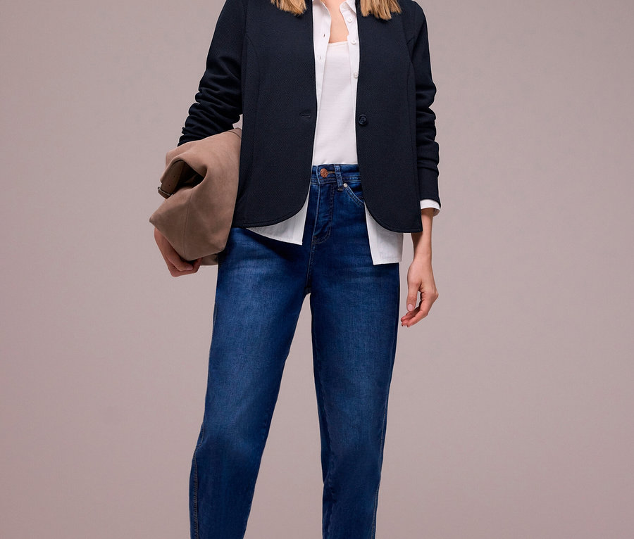 Lächelnde Frau trägt dunkelblauen Street One Jerseyblazer, weißes Oberteil, blaue Jeans und taupefarbene Ballerinas, eine braune Tasche haltend.