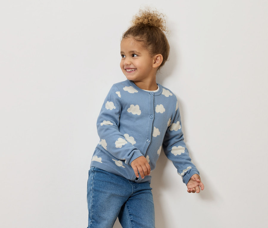 Ein lächelndes kleines Mädchen mit lockigem Haar trägt eine blaue Strickjacke mit Wolkenmuster und Jeans.