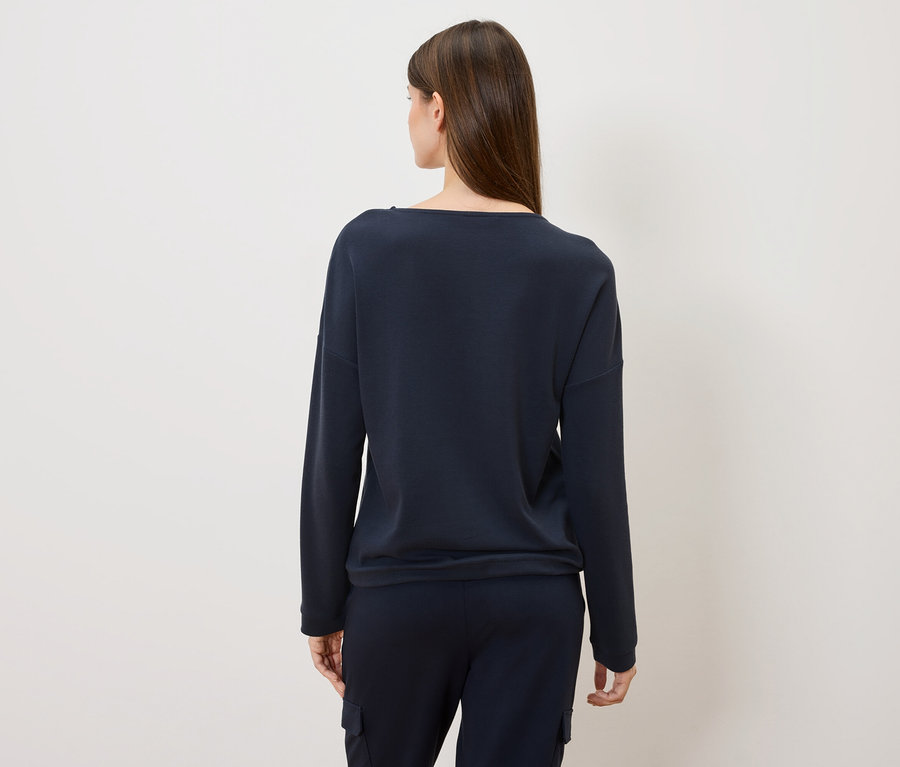 Rückansicht einer Frau mit langen braunen Haaren, die einen dunkelblauen Pullover und eine Hose trägt.