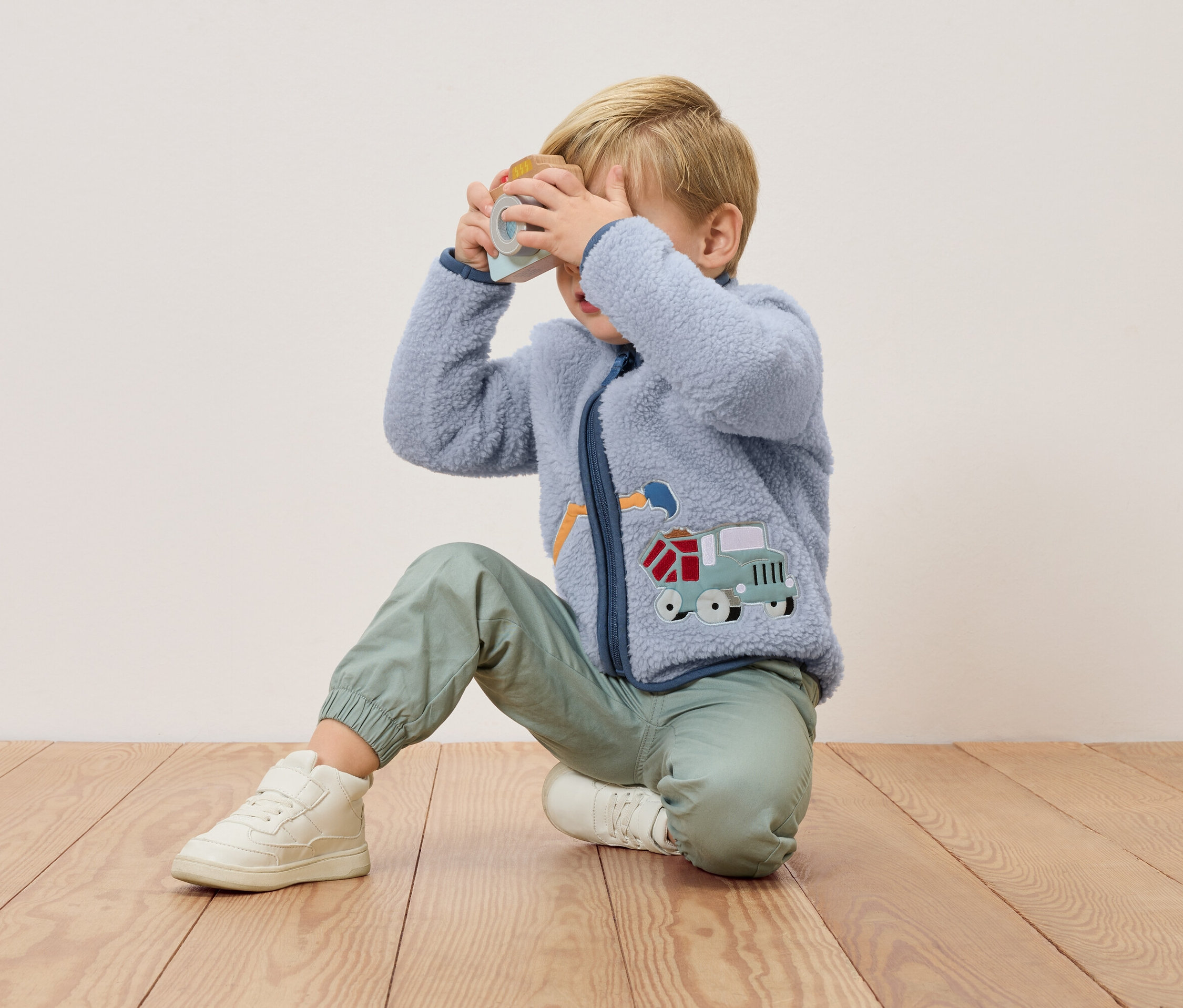 Ein blonder Junge kniet auf einem Holzboden und hält eine Spielzeugkamera vor sein Gesicht. Er trägt eine blaue Fleecejacke mit einem Lastwagen-Applikation und grüne Hosen.